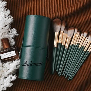 Puede incluir: Un juego de brochas de maquillaje con mangos verde oscuro y férulas doradas, junto con un estuche cilíndrico verde a juego. La palabra "Adomeit" está impresa en el estuche. También se ve un frasco de perfume con "Enjoy Life".