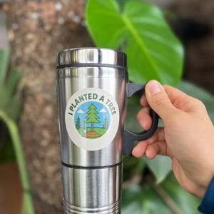 May include: A stainless steel travel mug with a black handle and lid. The mug features a circular sticker with the text "I PLANTED A TREE" and a graphic of a tree, grass, and a blue sky. The mug is held by a hand.