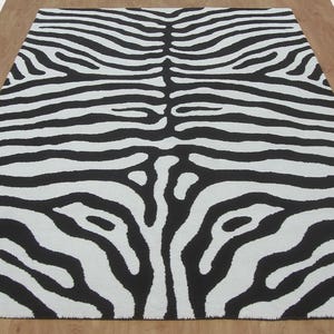 May include: A rectangular rug with a zebra stripe pattern in black and white. The rug is placed on a wooden floor, and the zebra stripes run horizontally across the rug. The rug is a decorative item for a living room or bedroom.
