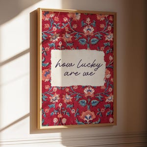 May include: A framed art print with a red floral pattern background. The print features a white rectangle with the text "how lucky are we" in a handwritten style. The frame is a light brown color.