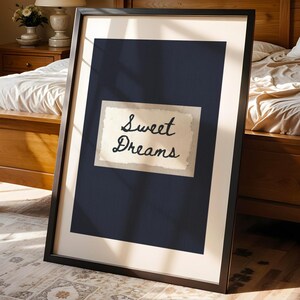 May include: A framed print with the words "Sweet Dreams" in elegant script. The artwork features a navy blue background with a cream-colored rectangle holding the text. The frame is black, and the print is leaning against a wooden bed frame.