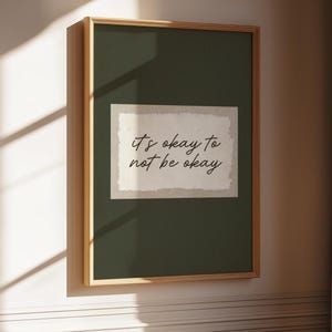 Puede incluir: Una impresión artística enmarcada con el texto "it's okay to not be okay" en un estilo manuscrito. La obra de arte presenta un fondo verde oscuro con un rectángulo de color claro en el centro. El marco es de color marrón claro.