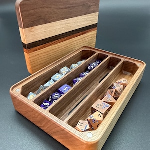May include: A rectangular wooden box with a removable lid, featuring three internal compartments. The box is crafted from light and dark wood strips. Inside are various multi-sided gaming dice in blue, purple, and copper colors with floral patterns.