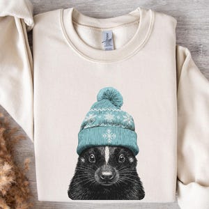 May include: A cream-coloured sweatshirt with a detailed illustration of a skunk wearing a light blue winter hat with snowflake patterns. The skunk's fur is black and white, with a focused gaze. The sweatshirt has a classic crew neck and long sleeves.