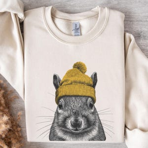 May include: Cream-coloured sweatshirt with a detailed illustration of a squirrel wearing a yellow knit hat with a pom-pom. The squirrel's face is in black and white, contrasting with the hat. The sweatshirt has a crew neck and long sleeves.