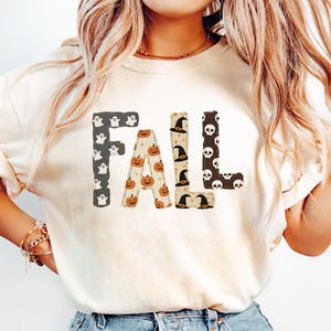 May include: A cream-colored short-sleeve t-shirt featuring the word FALL printed in large block letters. Each letter has a unique pattern: the F has white ghosts, the A has orange pumpkins, the L has witch hats, and the L has white skulls.