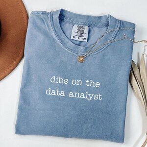 May include: A blue Comfort Colors t-shirt with the text "dibs on the data analyst" in white. A gold necklace with a pendant is draped across the shirt. The shirt is size small.