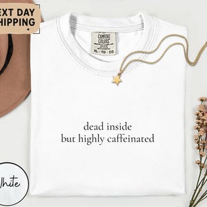 May include: A white Comfort Colors t-shirt with the text "dead inside but highly caffeinated" in black. A gold star pendant necklace rests on the shirt. A brown hat and flowers are in the background.