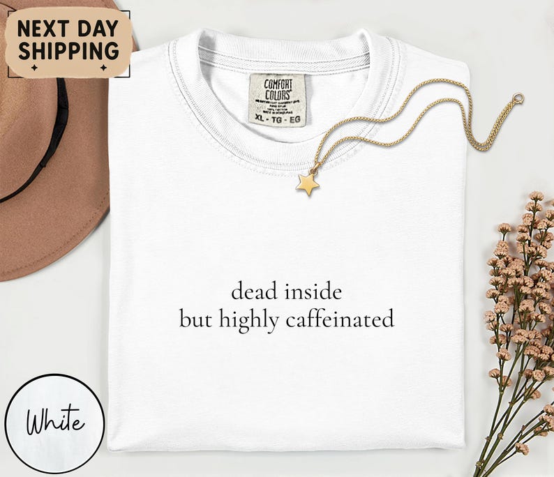 May include: A white Comfort Colors t-shirt with the text "dead inside but highly caffeinated" in black. A gold star pendant necklace rests on the shirt. A brown hat and flowers are in the background.