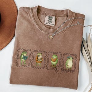 Puede incluir: Camiseta Comfort Colors marrón con un gráfico de cuatro ranas representadas como sellos postales, cada una leyendo un libro. Un collar plateado con un colgante descansa sobre la camiseta. La camiseta es talla S.