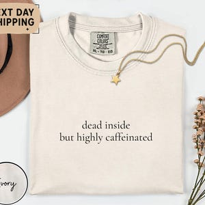 May include: Ivory-colored t-shirt with the text "dead inside but highly caffeinated." A gold star pendant necklace rests on the shirt. The shirt has a Comfort Colors tag. The image also includes a brown hat and floral arrangement.