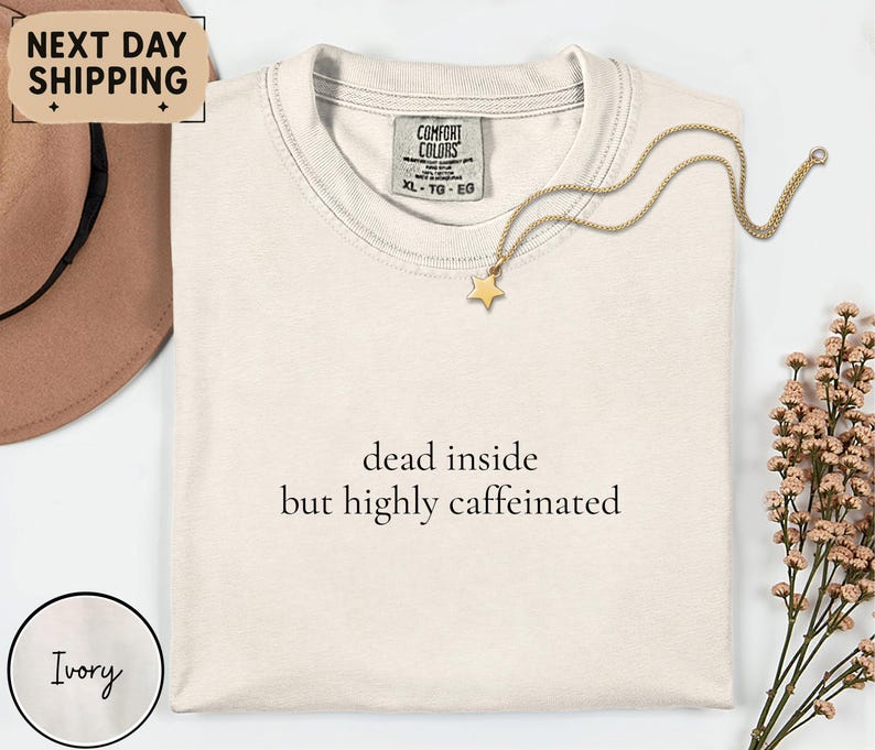 May include: Ivory-colored t-shirt with the text "dead inside but highly caffeinated." A gold star pendant necklace rests on the shirt. The shirt has a Comfort Colors tag. The image also includes a brown hat and floral arrangement.