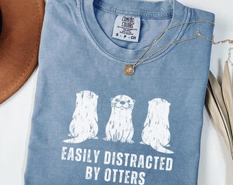 May include: A blue Comfort Colors t-shirt featuring the text "EASILY DISTRACTED BY OTTERS" and a graphic of three otters. A gold necklace with a pendant is draped across the shirt. The shirt is size small.