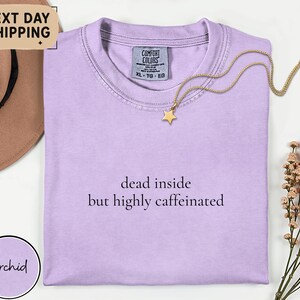 May include: A light purple t-shirt with the text "dead inside but highly caffeinated". A gold star pendant necklace rests on the shirt. The shirt is next to a brown hat and a sprig of dried flowers.