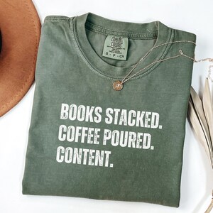 May include: An olive green t-shirt with the words "BOOKS STACKED. COFFEE POURED. CONTENT." in white block letters. A gold necklace with a pendant is draped across the shirt. The shirt is folded on a white surface.