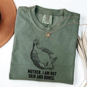 May include: Olive green t-shirt with a graphic of a duck and the text "MOTHER, I AM BUT SKIN AND BONES." A gold necklace with a pendant is draped across the shirt. The shirt is folded on a white surface.