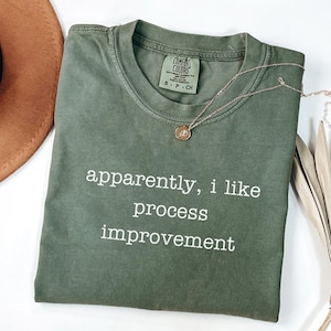 May include: An olive green t-shirt with the text "apparently, I like process improvement" in white. A gold necklace with a pendant is draped over the shirt. The shirt is folded on a white surface.