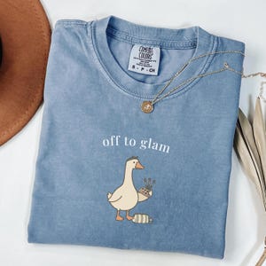 Pode incluir: Uma camiseta azul Comfort Colors com a frase "off to glam" e um desenho de um ganso com pincéis de maquiagem. Um colar dourado e um chapéu marrom também são visíveis.