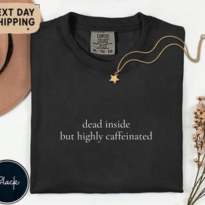 May include: Black Comfort Colors t-shirt with the text "dead inside but highly caffeinated" in white. A gold star pendant necklace is draped on the shirt. A brown hat and a sprig of flowers are in the background.