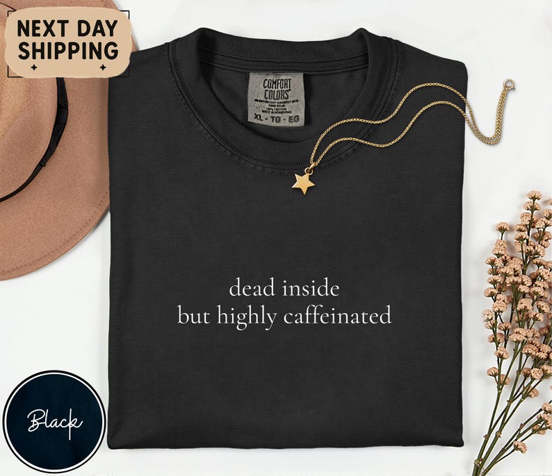 May include: Black Comfort Colors t-shirt with the text "dead inside but highly caffeinated" in white. A gold star pendant necklace is draped on the shirt. A brown hat and a sprig of flowers are in the background.