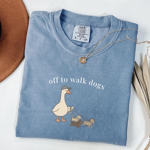 Peut inclure: T-shirt bleu clair Comfort Colors avec l'inscription "off to walk dogs" et une oie de dessin animé portant un chapeau et promenant trois petits chiens. Un collier en or est drapé sur le t-shirt.