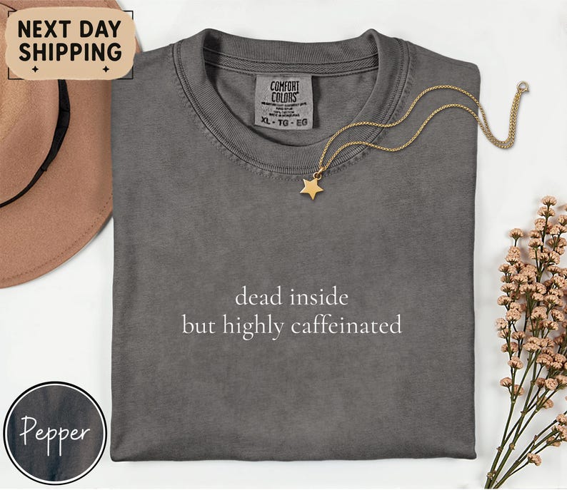 May include: A gray Comfort Colors t-shirt with the text "dead inside but highly caffeinated" in white. A gold star pendant necklace is draped across the shirt. A brown hat and a sprig of pink flowers are in the background.