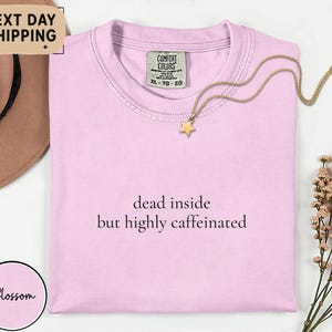 May include: A light pink t-shirt with the text "dead inside but highly caffeinated" in black. A gold star pendant necklace rests on the shirt. The shirt has a Comfort Colors tag.