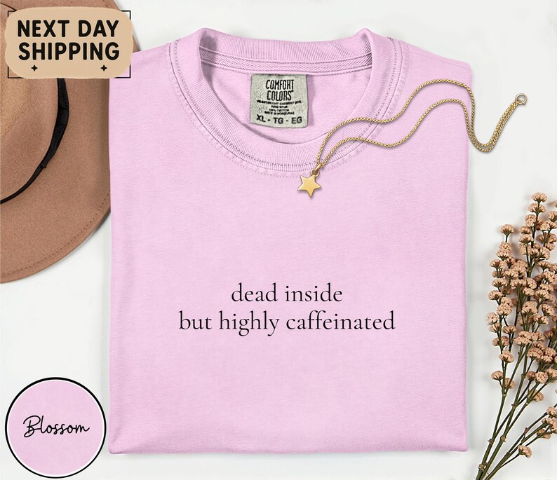 May include: A light pink t-shirt with the text "dead inside but highly caffeinated" in black. A gold star pendant necklace rests on the shirt. The shirt has a Comfort Colors tag.
