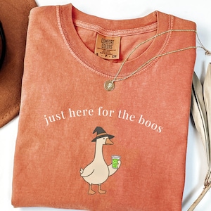 May include: A terracotta-colored crew neck t-shirt featuring a graphic of a goose wearing a black witch hat and holding a green drink. The text "just here for the boos" is printed in white above the goose. The shirt has a Comfort Colors label.