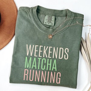 May include: An olive green t-shirt with the words "WEEKENDS MATCHA RUNNING" printed in white, green, and pink. A gold necklace with a pendant is draped over the shirt. The shirt is folded on a white surface.