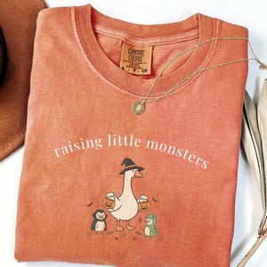 Pode incluir: Uma t-shirt de gola redonda cor de terracota com o texto branco "raising little monsters" sobre um gráfico de um ganso com um chapéu de bruxa que segura dois baldes de abóbora, ladeado por um pequeno pinguim e uma personagem de dinossauro.