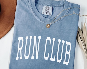 May include: A blue t-shirt with the words "RUN CLUB" in white block letters. A gold necklace with a round pendant is draped across the shirt. The shirt is a Comfort Colors brand, size S-P-CH.