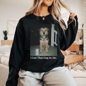 May include: Black sweatshirt featuring a framed image of a dog with a cigarette in its mouth and a can. The text "I Got That Dog In Me" is below the image. The dog is brown and gray.