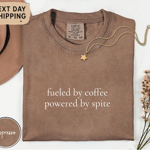 以下が含まれることがあります： 「fueled by coffee powered by spite」の文字が入ったエスプレッソカラーのTシャツ。ゴールドの星型ペンダントがゴールドチェーンにぶら下がっています。シャツは折りたたまれており、Comfort Colorsのタグが付いています。茶色の帽子と花が背景にあります。