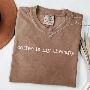 May include: A brown Comfort Colors t-shirt with the phrase "coffee is my therapy" in white lettering. A silver necklace with a small pendant rests on the shirt. A brown hat is visible in the upper left corner.