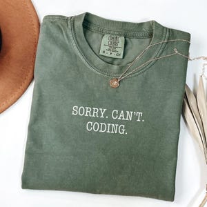 Op de afbeelding: Olijfgroen T-shirt met de witte tekst "SORRY. CAN'T. CODING.". Een goudkleurige ketting met een kleine hanger ligt over het shirt. Het shirt is opgevouwen en op een wit oppervlak geplaatst.