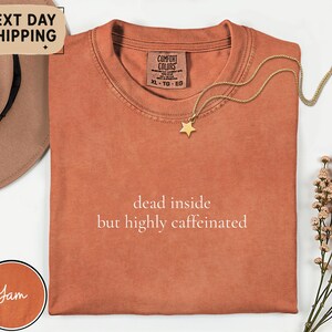 May include: A terracotta-colored t-shirt with the text "dead inside but highly caffeinated" in white. A gold star pendant necklace rests on the shirt. The shirt is folded, and a brown hat is partially visible.