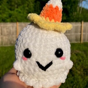 May include: A white crocheted ghost with black eyes, pink cheeks, and a black smile. The ghost wears a candy corn hat with orange, yellow, and white colours. The plush toy is held in a hand against a blurred green background.