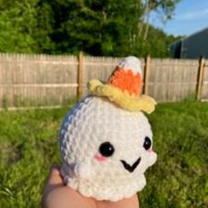 Op de afbeelding: Een handgemaakt wit gehaakt spook met zwarte ogen, een lachende mond en roze wangen. Het spook draagt een kleine, gele hoed met een snoepma&iuml;smotief. Het spook wordt in de hand van een persoon gehouden tegen een achtergrond van groen gras en een houten hek.