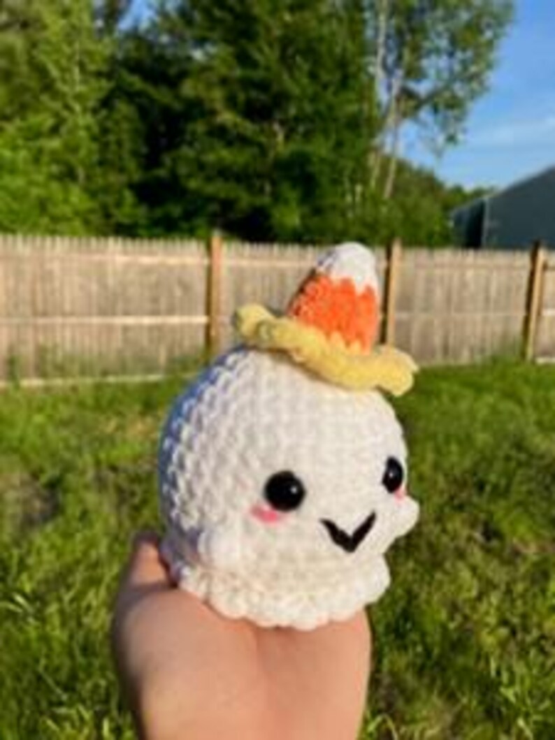 Op de afbeelding: Een handgemaakt wit gehaakt spook met zwarte ogen, een lachende mond en roze wangen. Het spook draagt een kleine, gele hoed met een snoepma&iuml;smotief. Het spook wordt in de hand van een persoon gehouden tegen een achtergrond van groen gras en een houten hek.