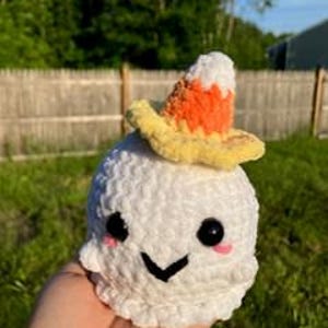 Op de afbeelding: Een witte gehaakte geest met een lachend gezicht en een snoepma&iuml;shoed. De geest heeft zwarte ogen, roze wangen en een zwart gestikte mond. De hoed is geel, oranje en wit. De geest is handgemaakt.