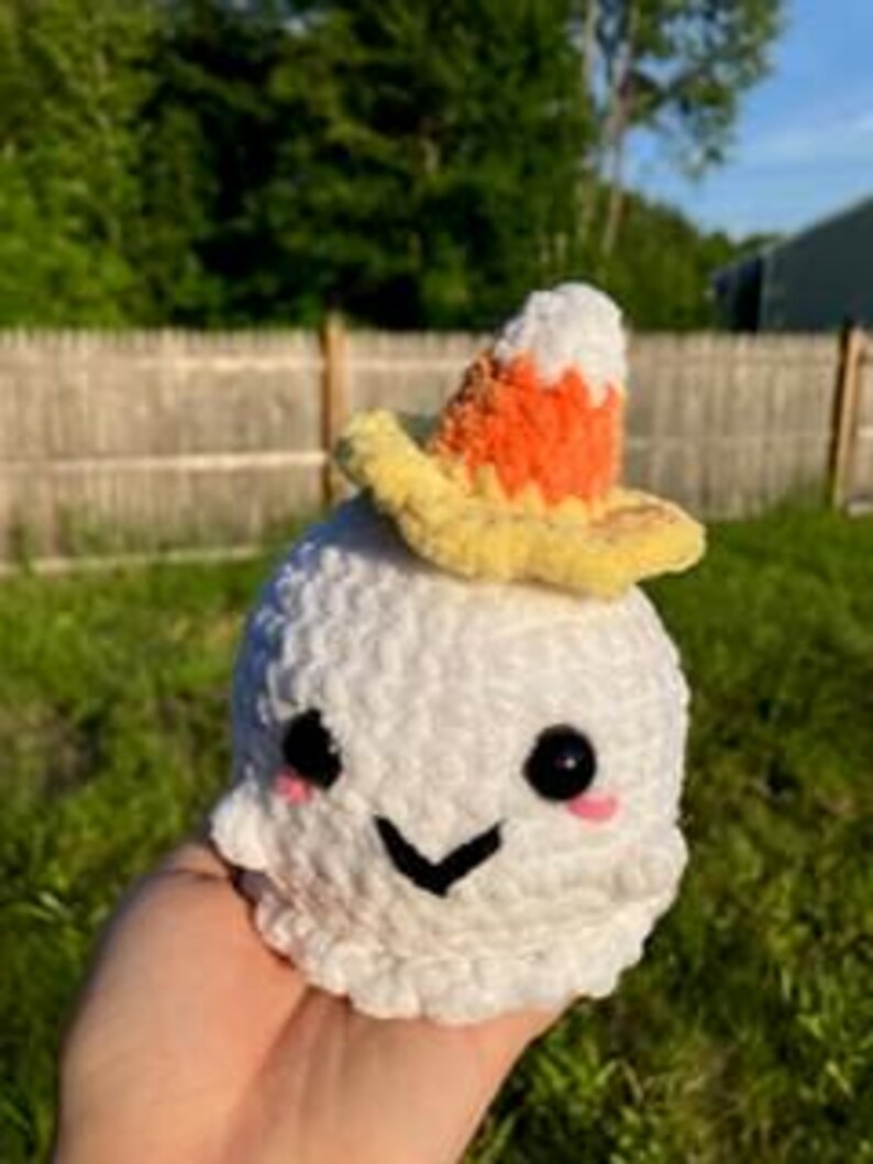 Op de afbeelding: Een witte gehaakte geest met een lachend gezicht en een snoepma&iuml;shoed. De geest heeft zwarte ogen, roze wangen en een zwart gestikte mond. De hoed is geel, oranje en wit. De geest is handgemaakt.