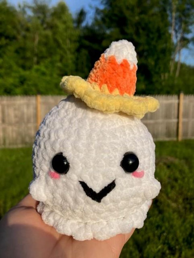 Op de afbeelding: Een wit gehaakt spook met zwarte ogen, roze wangen en een zwarte glimlach. Het spook draagt een snoepma&iuml;shoed met oranje, gele en witte kleuren. Het pluche speelgoed wordt in een hand gehouden tegen een vage groene achtergrond.