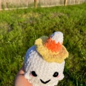 Op de afbeelding: Een kleine, witte gehaakte geest met een lachend gezicht en zwarte ogen. De geest draagt een hoed in de vorm van een snoepma&iuml;s met gele, oranje en witte strepen. De geest wordt in een hand gehouden tegen een achtergrond van groen gras en een houten hek.