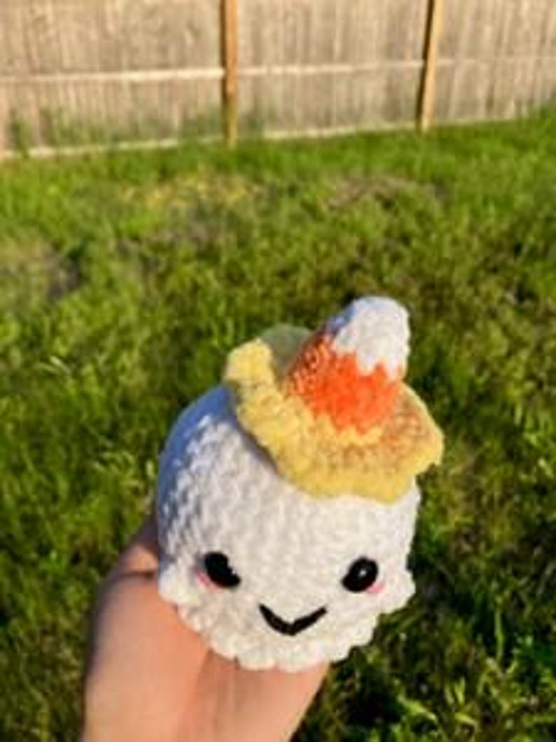 Op de afbeelding: Een kleine, witte gehaakte geest met een lachend gezicht en zwarte ogen. De geest draagt een hoed in de vorm van een snoepma&iuml;s met gele, oranje en witte strepen. De geest wordt in een hand gehouden tegen een achtergrond van groen gras en een houten hek.