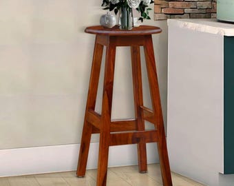 May include: A tall, wooden bar stool with a round seat and four angled legs. The stool is made of a rich, reddish-brown wood and has a simple, classic design. A vase of white flowers sits on top of the stool.