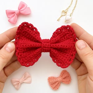 Pu&ograve; includere: Un fiocco all'uncinetto rosso brillante con bordo smerlato. Il fiocco &egrave; centrato, affiancato da un piccolo fiocco in raso rosa, una collana color oro con ciondoli a forma di chiave e cuore, un piccolo fiocco rosa chiaro e un piccolo fiocco all'uncinetto color pesca su una superficie bianca.