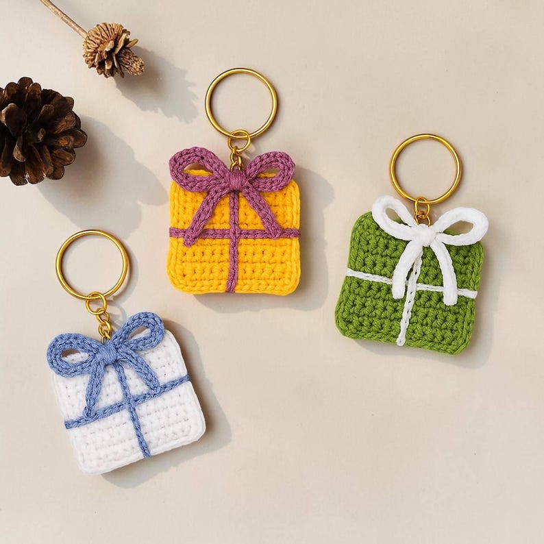 Peut inclure: Trois porte-cl&eacute;s faits &agrave; la main en forme de bo&icirc;te cadeau au crochet, de couleurs jaune, vert et blanc. Chaque bo&icirc;te est orn&eacute;e d'un n&oelig;ud sur le dessus et d'anneaux dor&eacute;s. La bo&icirc;te jaune a un n&oelig;ud violet, la bo&icirc;te verte a un n&oelig;ud blanc et la bo&icirc;te blanche a un n&oelig;ud bleu.
