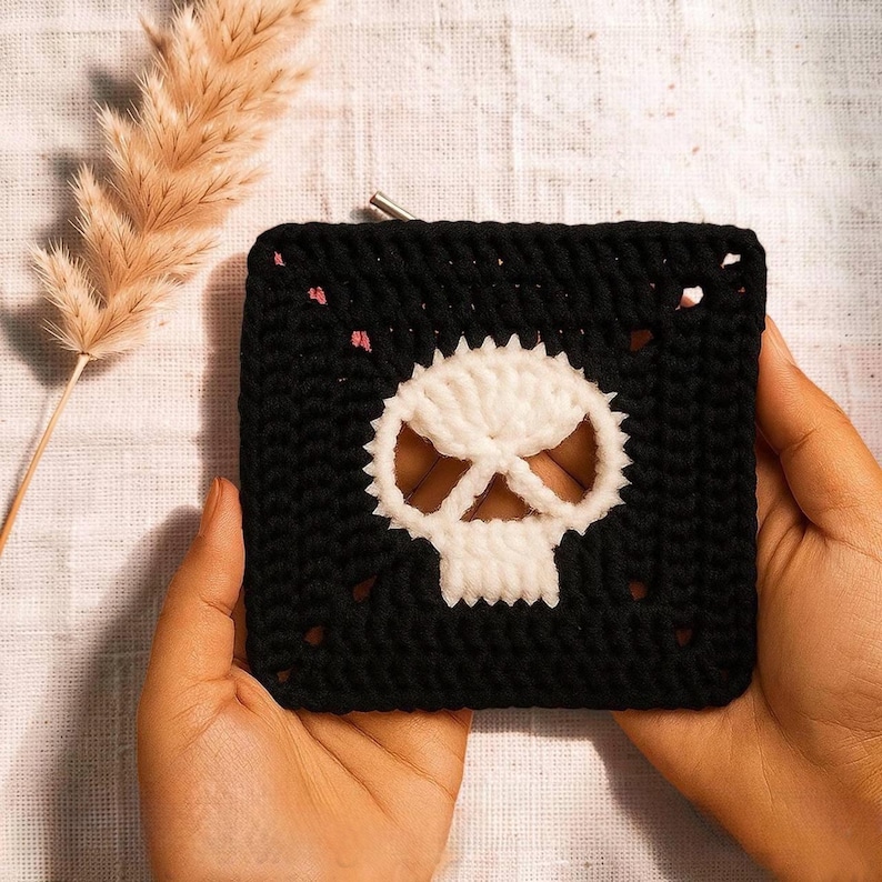 May include: A square crochet pouch made of black yarn featuring a white skull motif centered on the front. The pouch has a metallic zipper closure along the top edge and a textured, hand-stitched appearance with visible yarn loops.