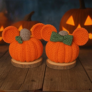 May include: Two crocheted orange pumpkins with round ears, each resting on a circular wood slice base. One pumpkin features a small green leaf and a gray stem, while the other displays a green bow and a gray stem, both crafted with yarn.